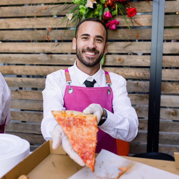 Smiling Pino's La Forchetta server in a pink apron offering a slice of cheese pizza.